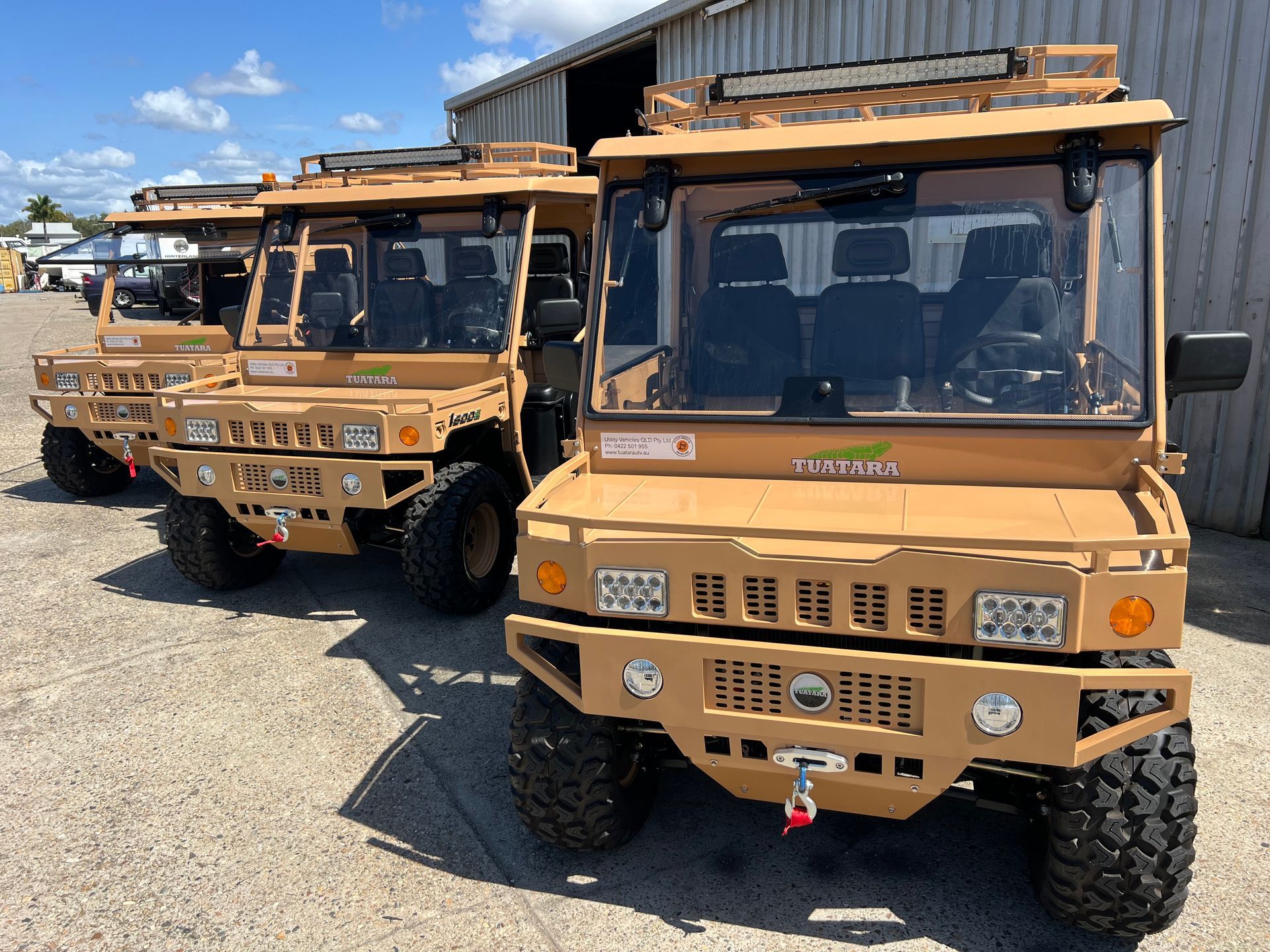 Three tan, off-road utility vehicles parked in a row outside a corrugated metal building on a sunny day.