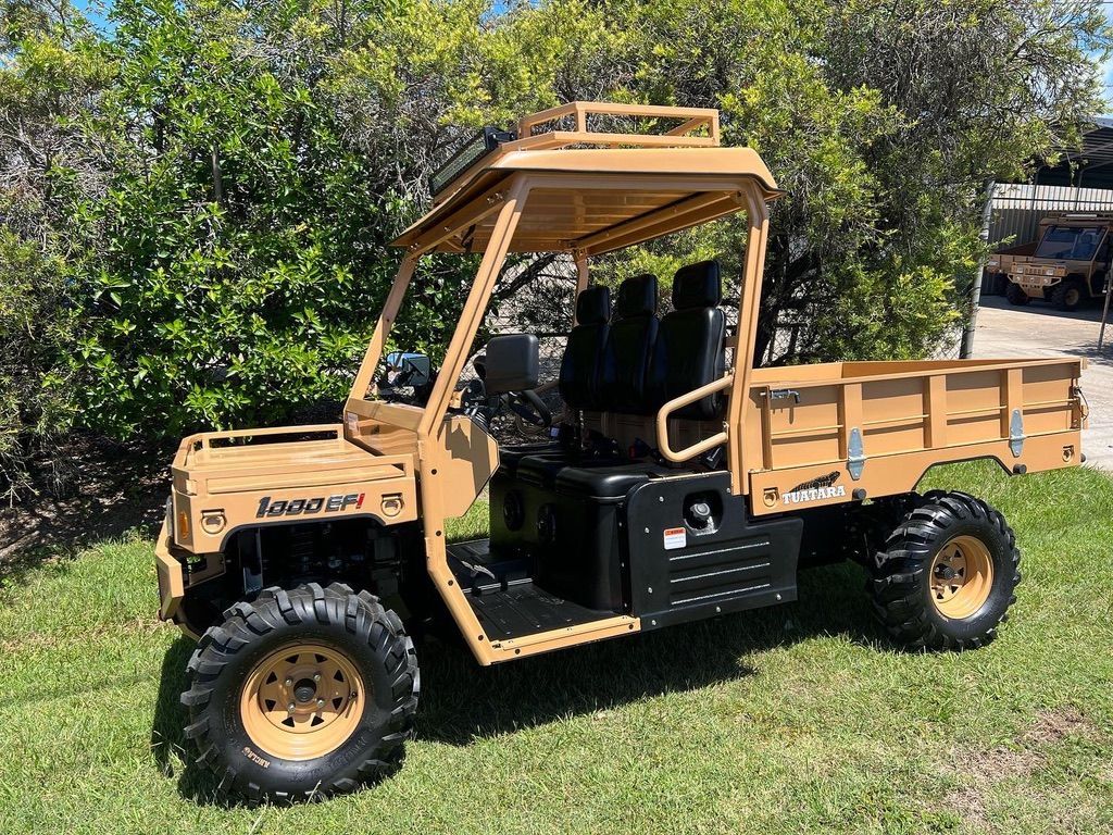 Tan, heavy-duty utility vehicle parked on grass in front of trees, featuring a flatbed and a roof-mounted light bar.