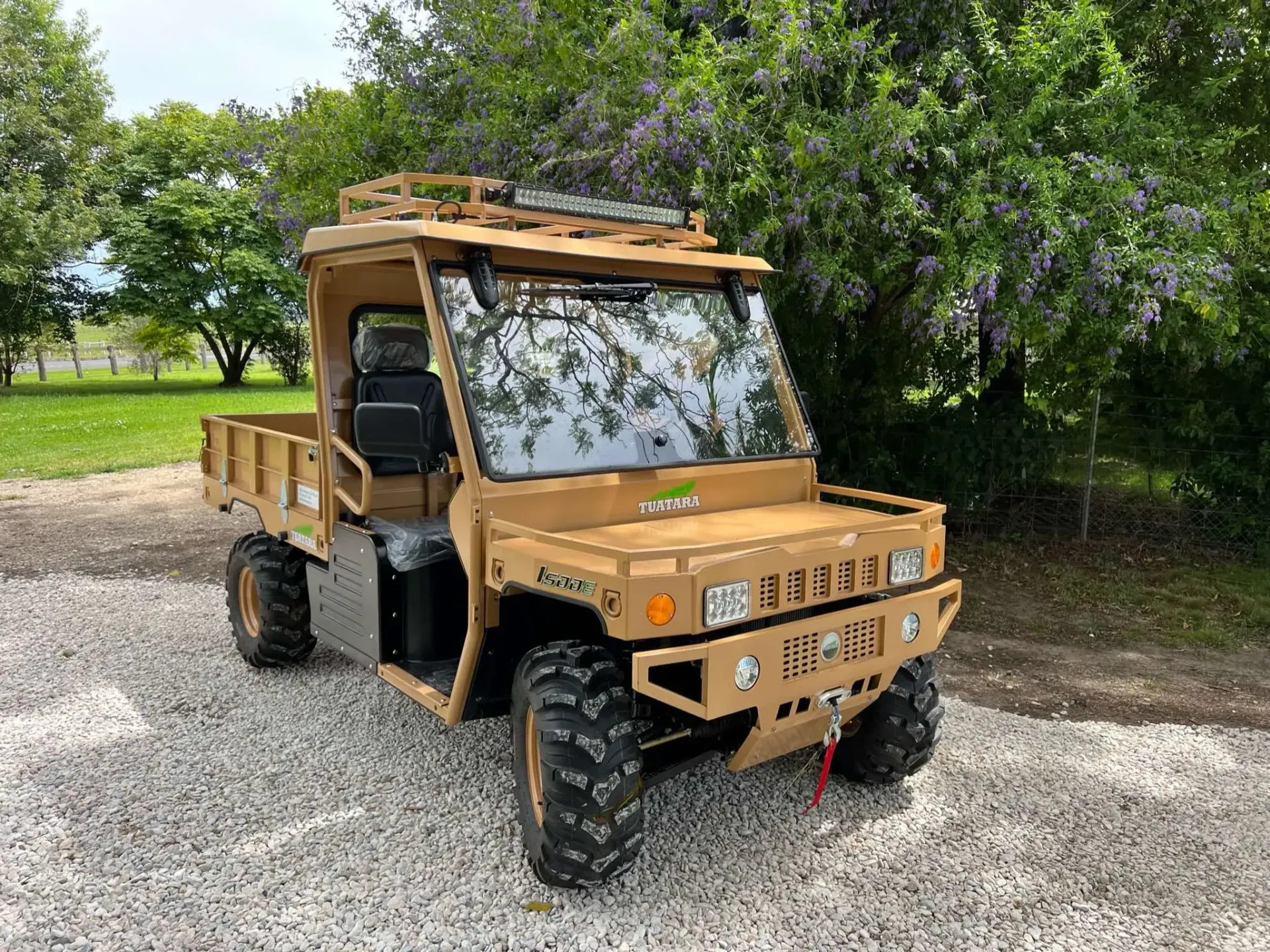 A tan, rugged utility terrain vehicle (UTV) parked on a gravel path outdoors with trees in the background.