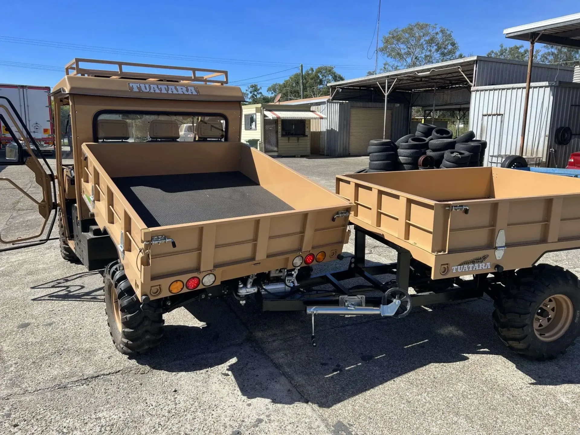 A tan utility vehicle with a dump bed attached to a matching cargo trailer parked on a gravel lot under a clear blue sky.