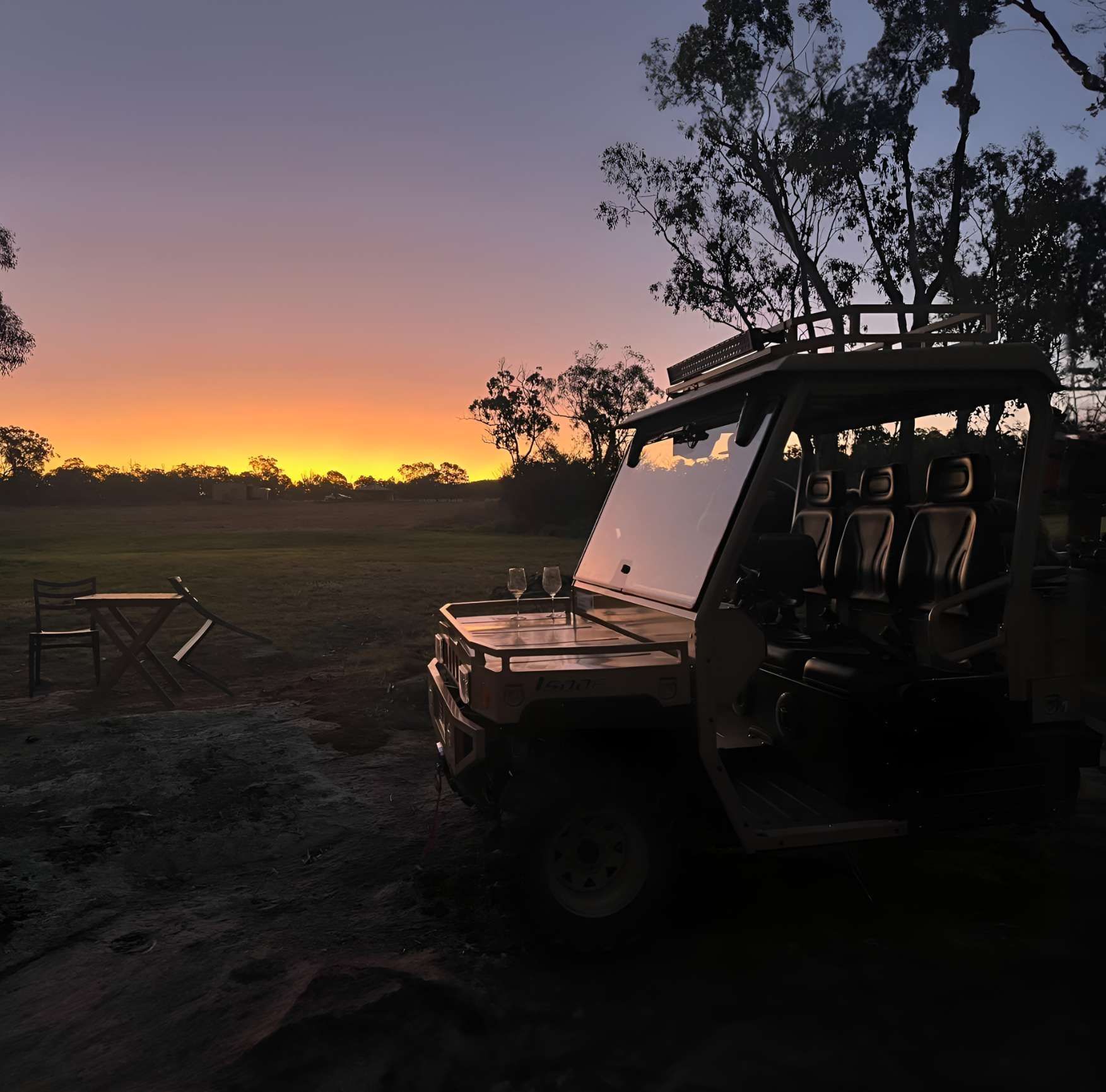 A safari vehicle parked in a field during a vibrant, colorful sunset with a small table nearby.