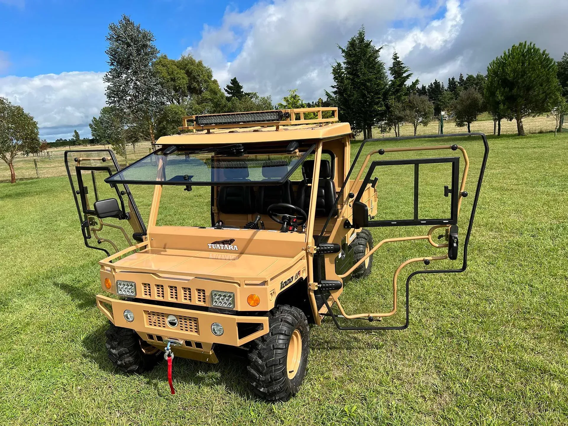 A tan, military-style utility vehicle with open doors, parked on a grassy field under a bright, cloudy sky.