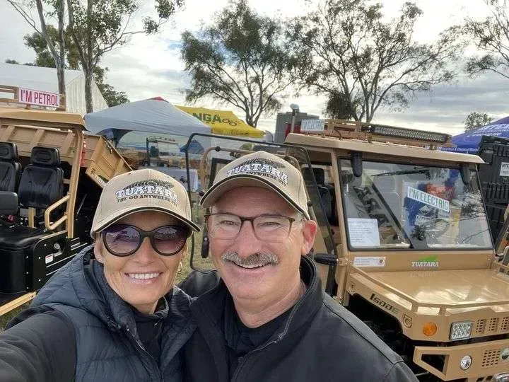 A couple in matching baseball caps smiles in a selfie outdoors, positioned in front of tan utility vehicles at an event.