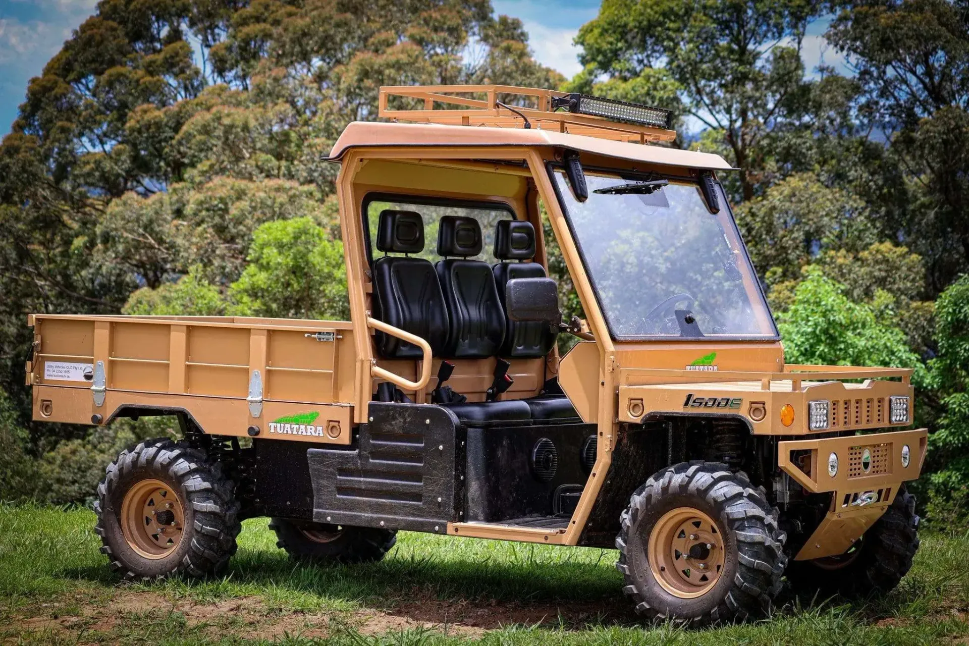 A tan, utilitarian off-road utility vehicle with a flatbed, parked in a grassy area with trees in the background.