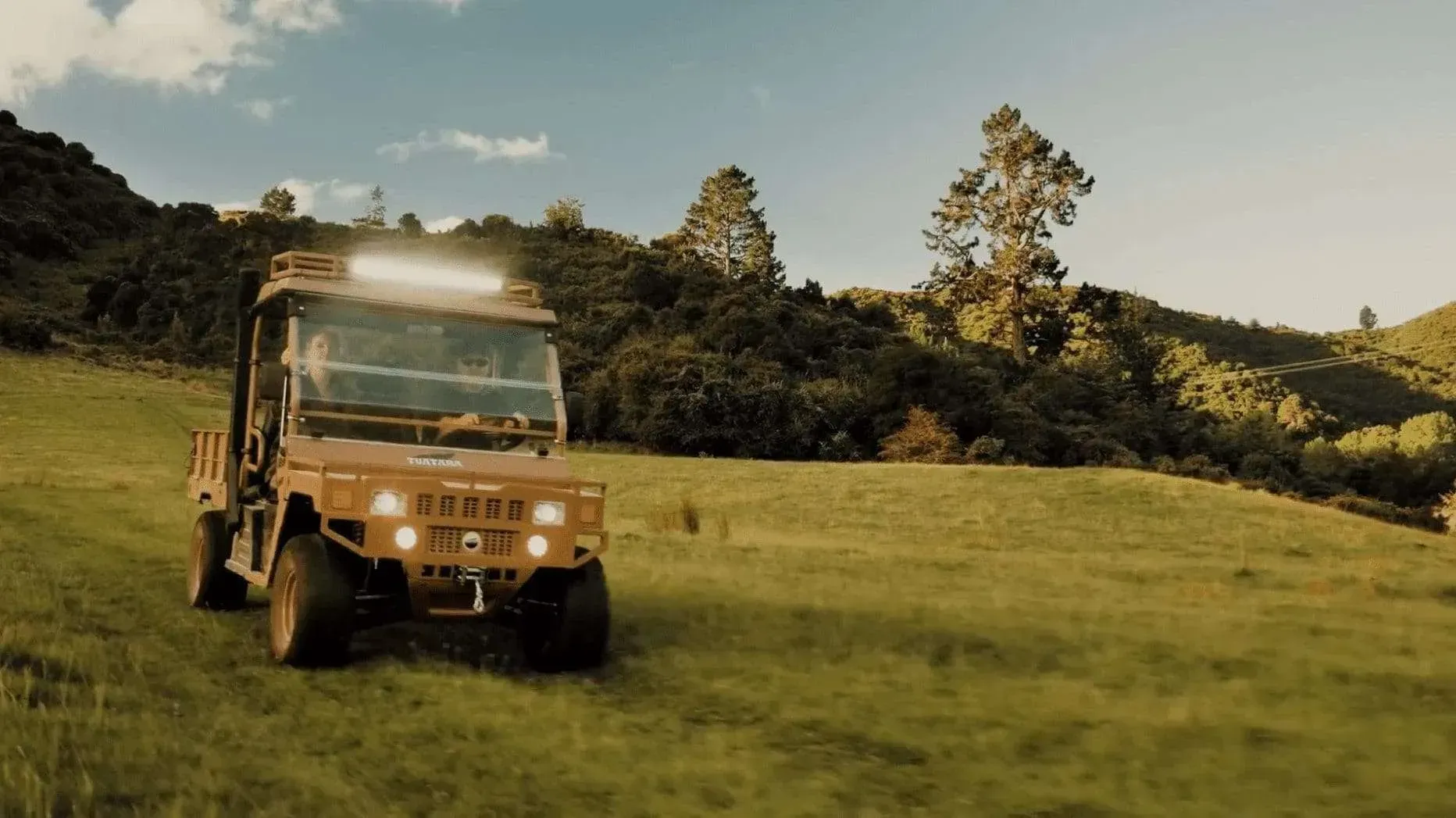 An orange utility terrain vehicle (UTV) drives across a green, grassy hillside under a clear blue sky.