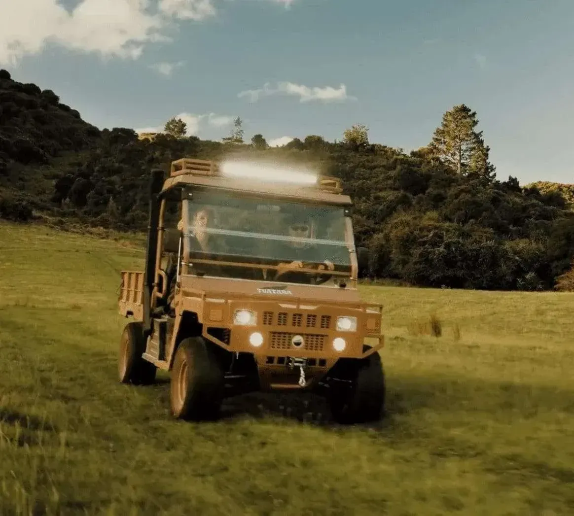 A tan utility terrain vehicle drives across a grassy field under a bright, partly cloudy sky.