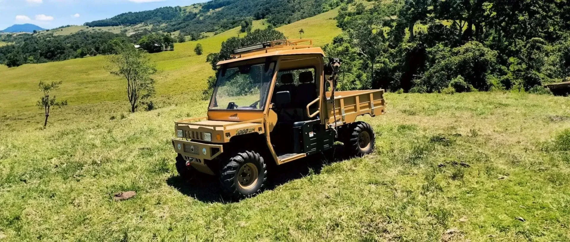 A tan utility terrain vehicle (UTV) parked on a grassy hillside under a clear blue sky.