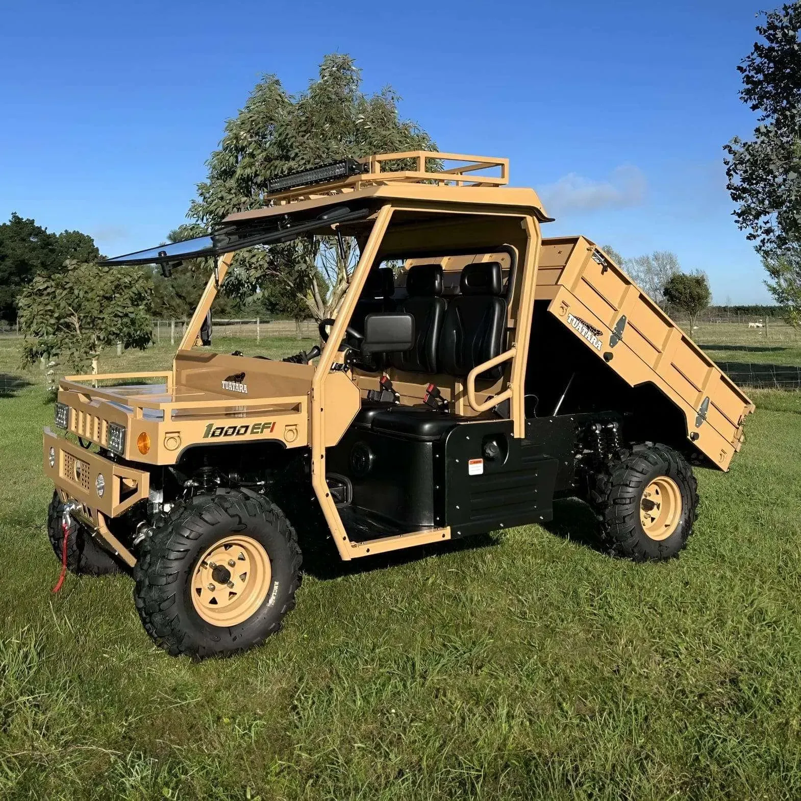 A tan utility task vehicle with a dump bed and roll cage parked in a grassy field under a clear blue sky.