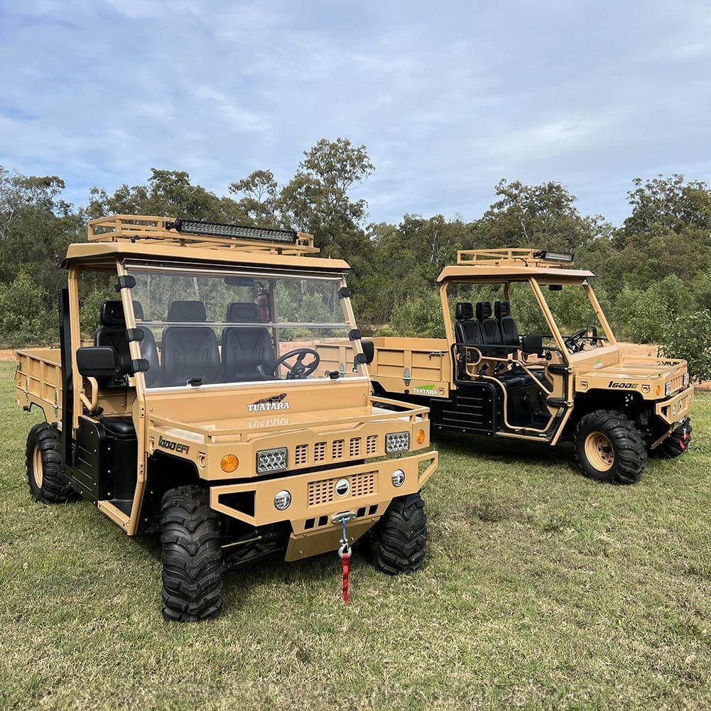 Two tan, rugged utility task vehicles parked on a grassy field with trees in the background under a blue sky.