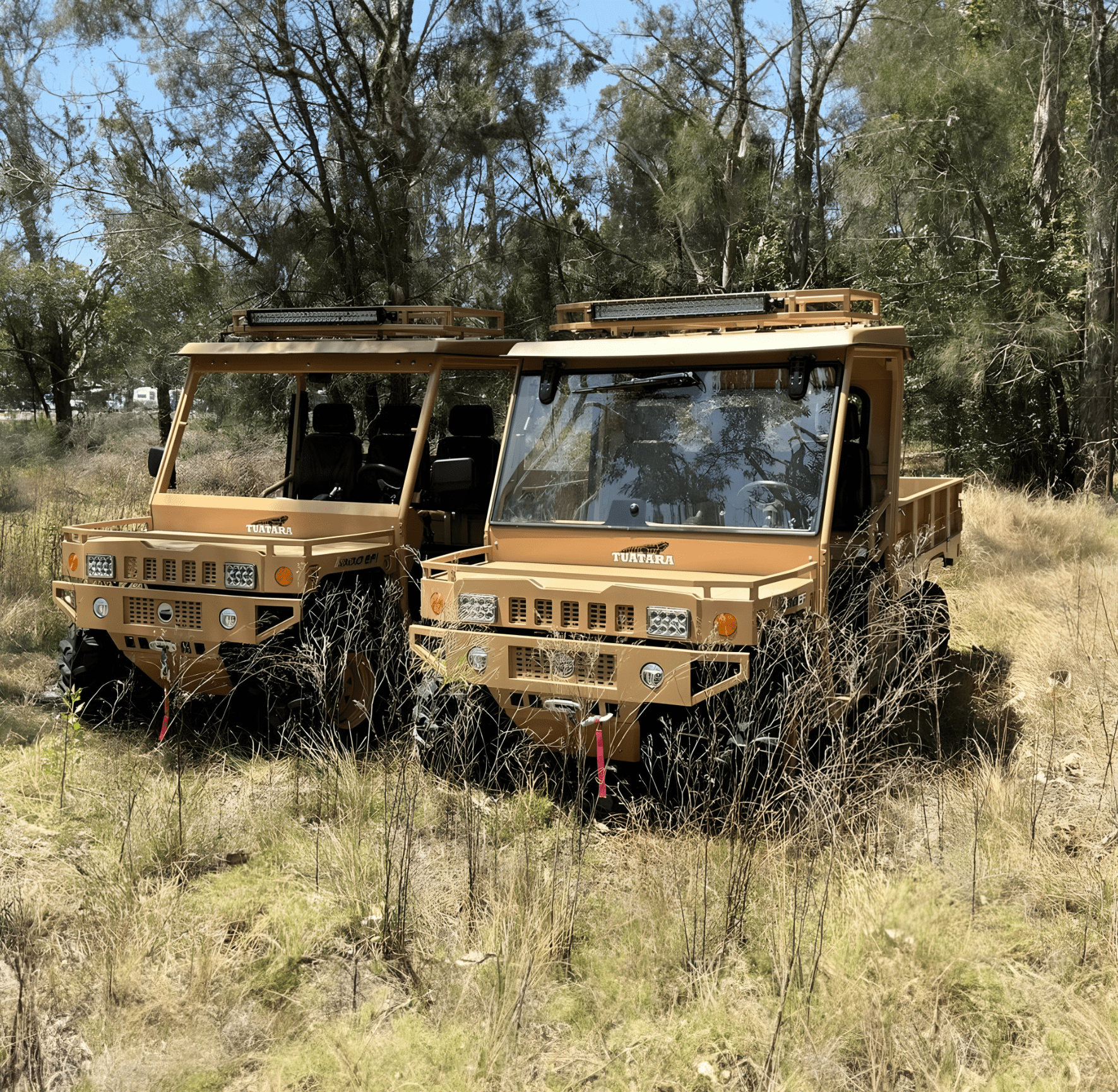 Two tan, military-style utility vehicles parked in a grassy, wooded field.