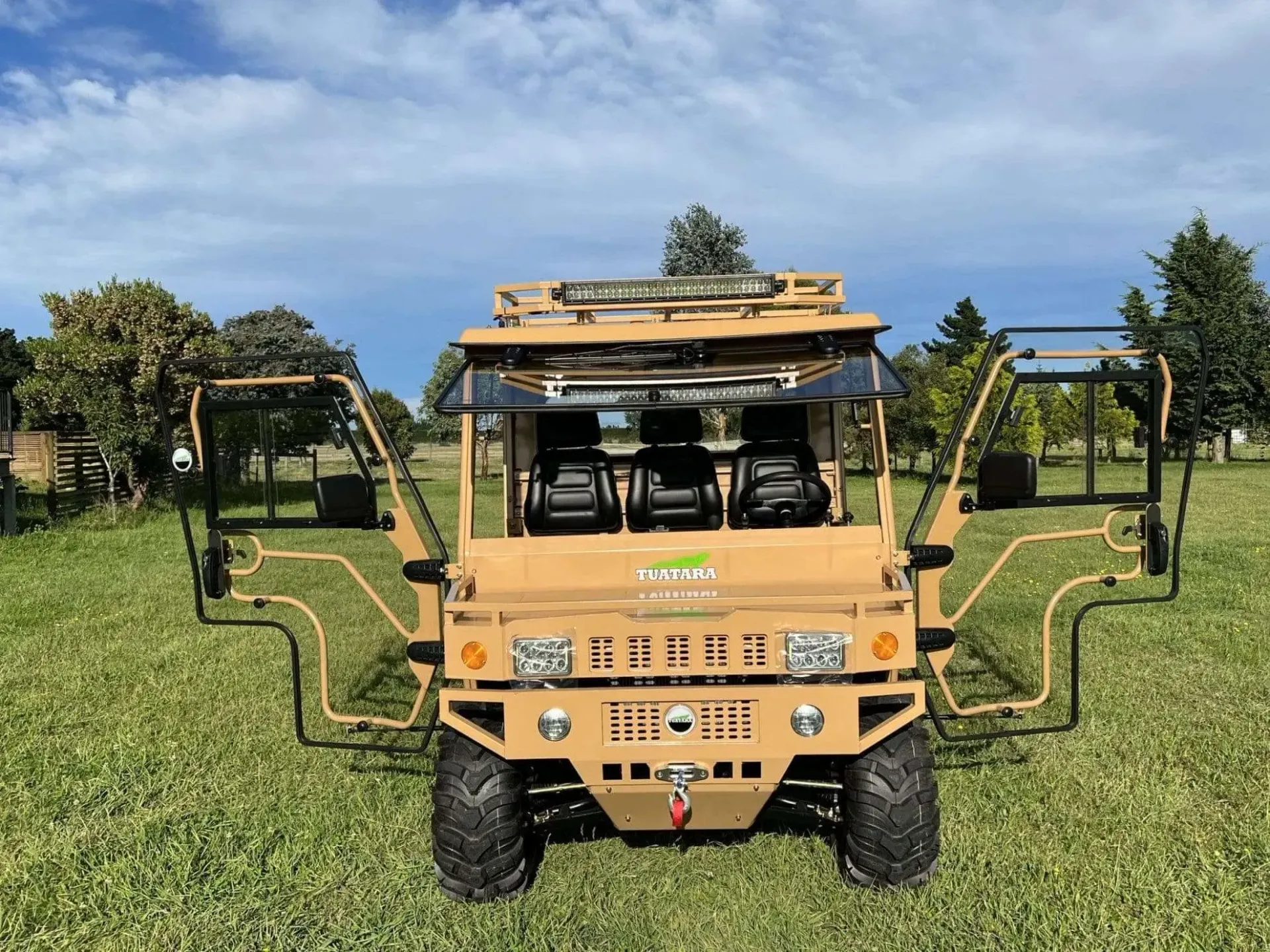 A tan military-style utility vehicle with open doors, parked in a grassy field under a sunny blue sky.