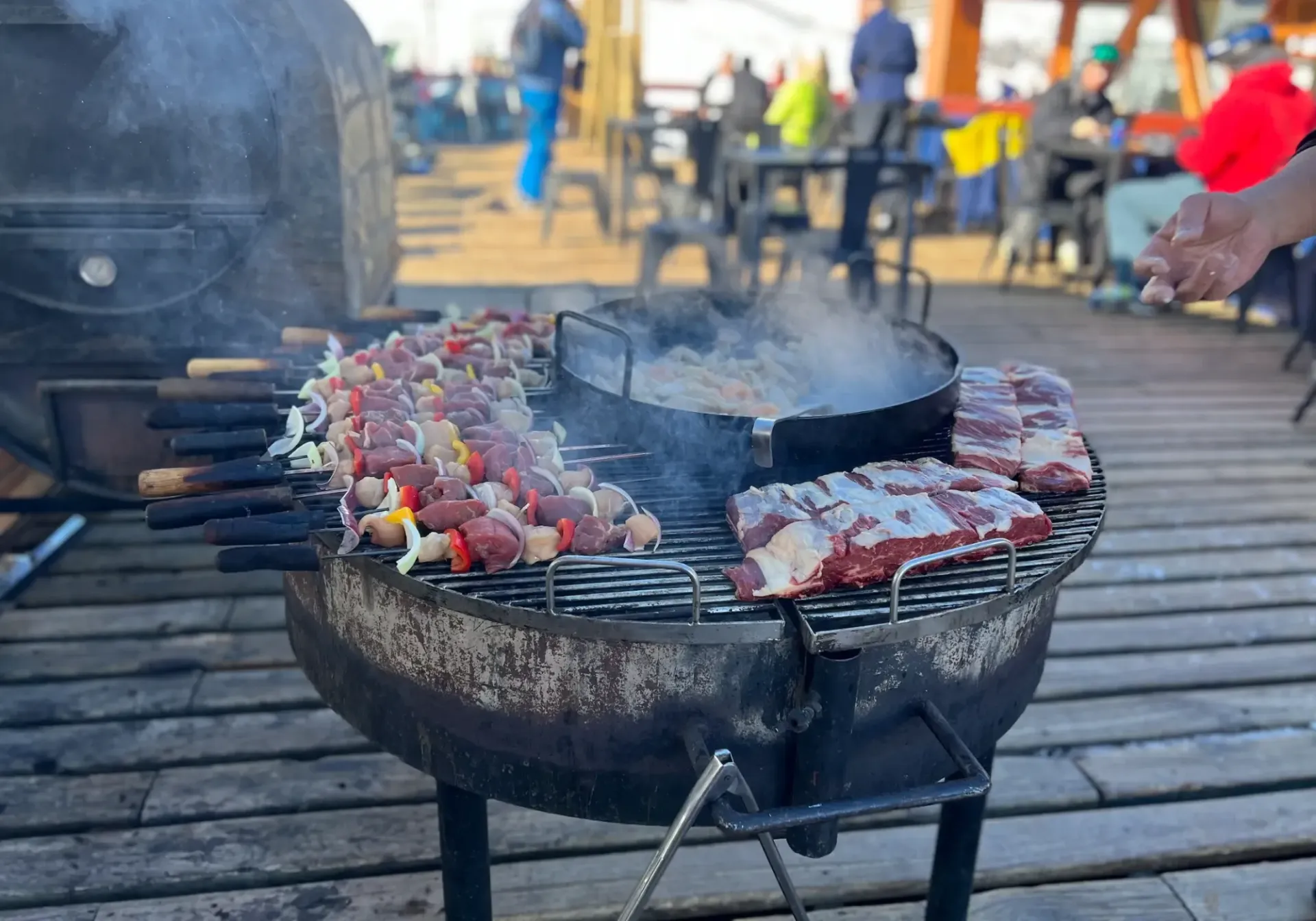 Skewers and meat cooking on a round grill with smoke rising outdoors on a deck