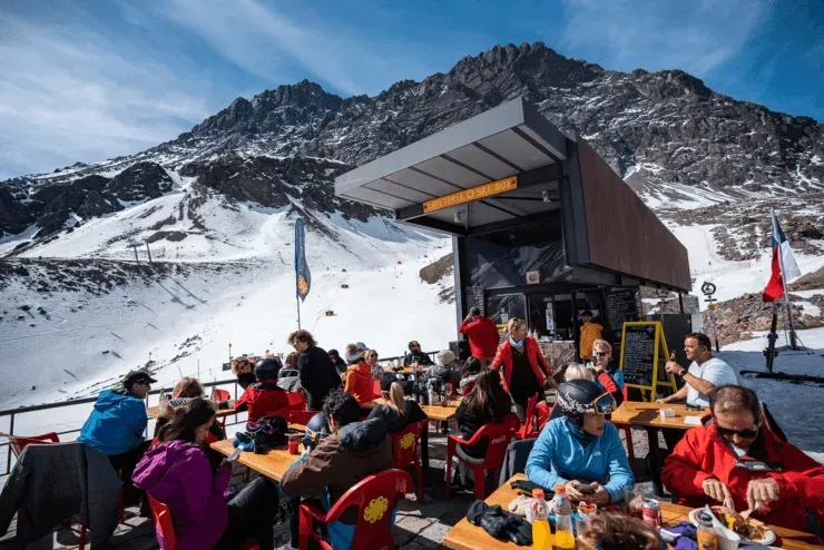 People dining at an outdoor ski lodge terrace with snowy mountains in the background