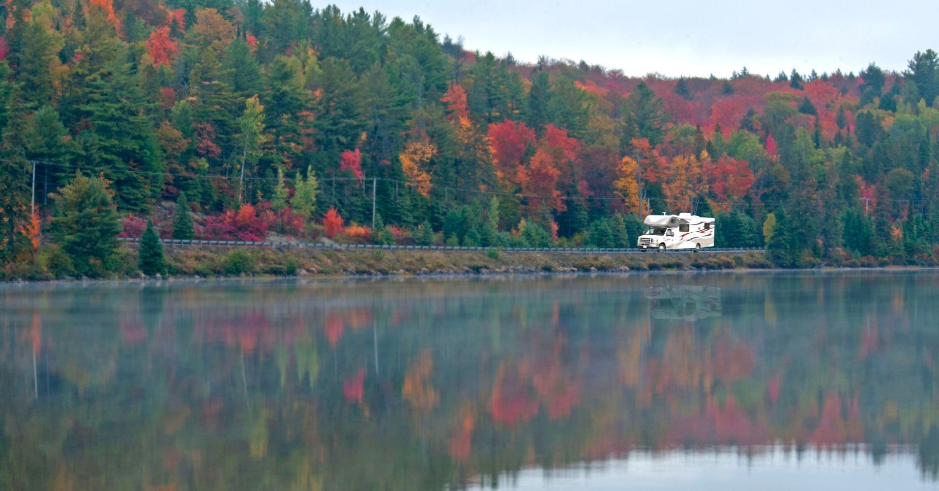 A rv is parked on the shore of a lake in the woods.