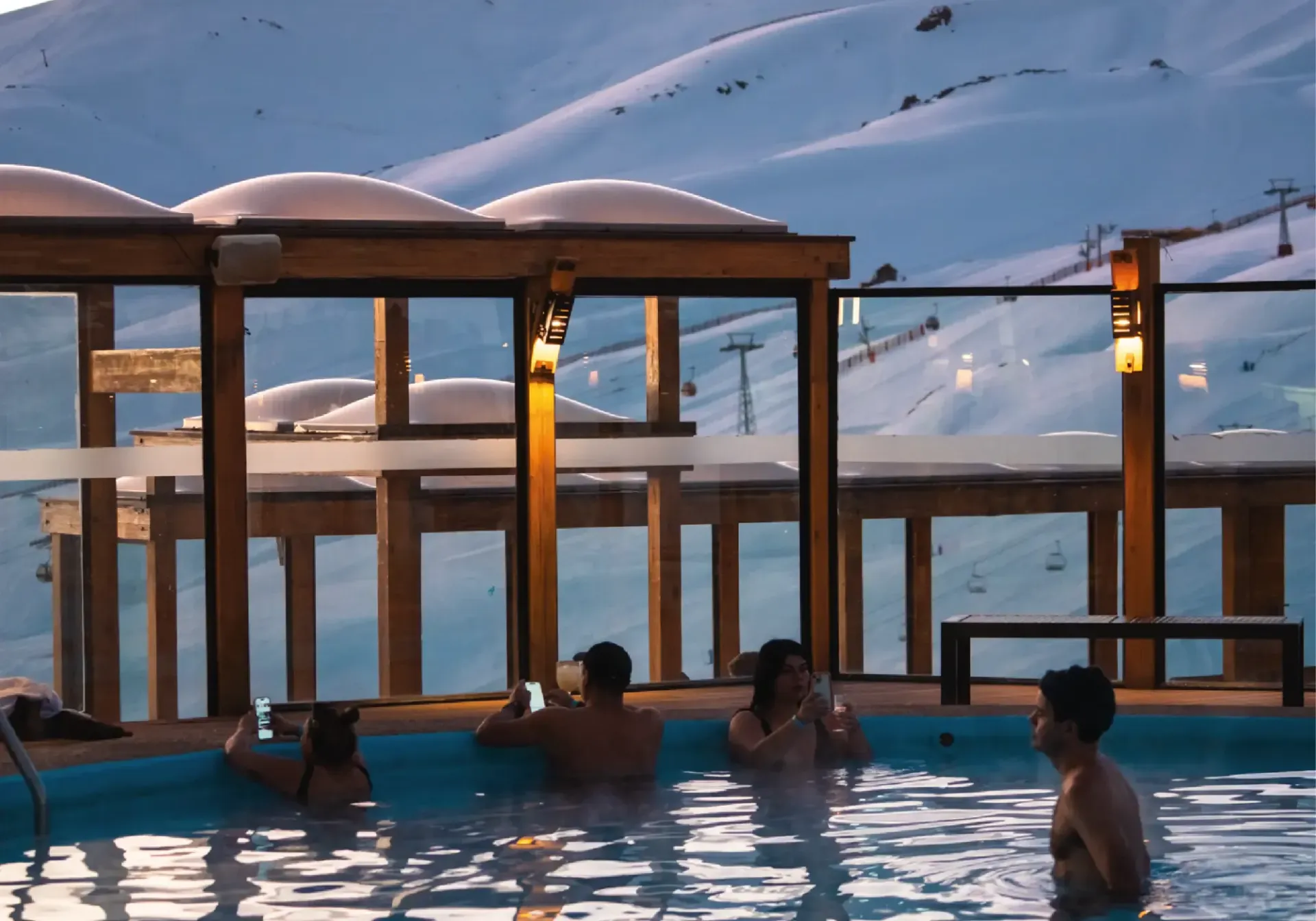 People relaxing in a rooftop pool at dusk, with mountains and snow outside the glass enclosure.
