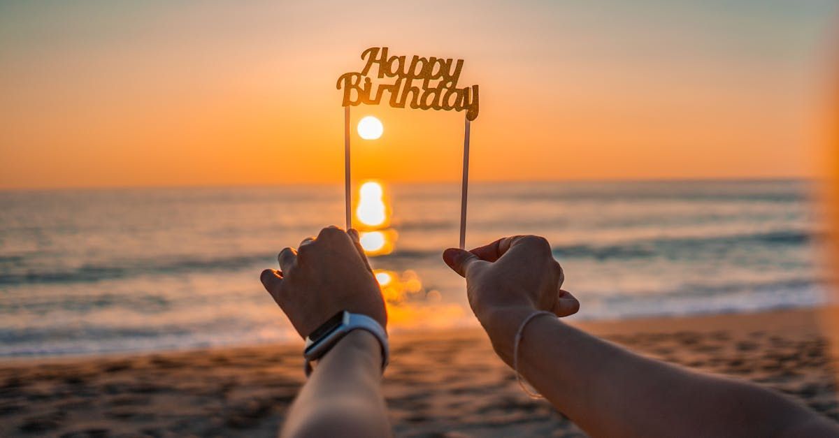 A person is holding a happy birthday cake topper on the beach.