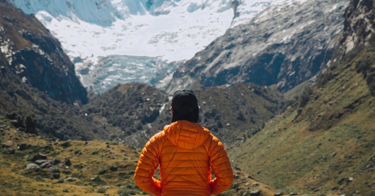 A person in an orange jacket is standing in front of a snowy mountain.