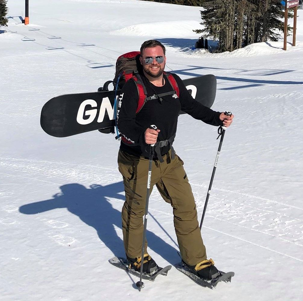 Man with backpack and ski poles standing on snowshoes on a snowy slope, holding a snowboard