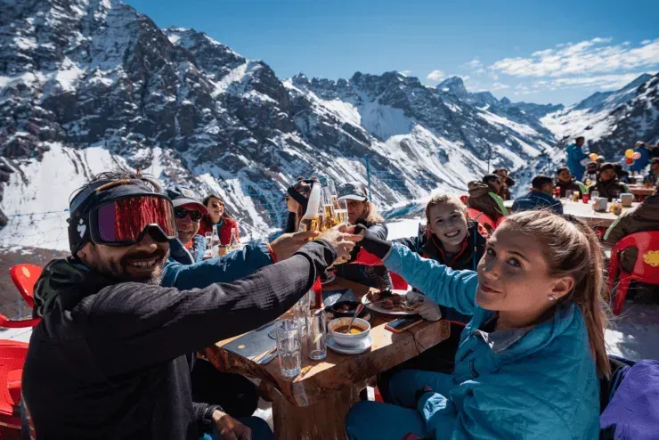 Skiers in colorful gear toast drinks at a snowy mountain lodge with dramatic peaks behind them