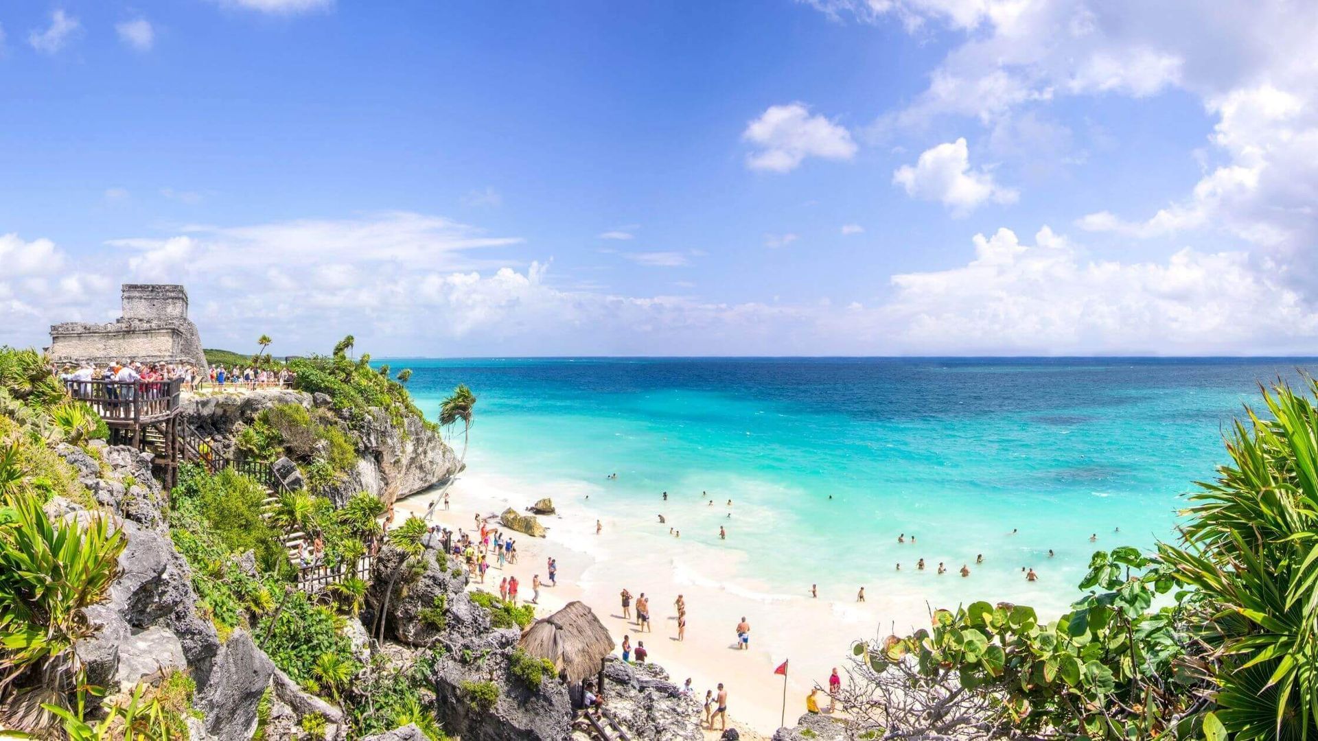 Sunlit tropical beach with turquoise water, white sand, cliffs, and swimmers under a blue sky