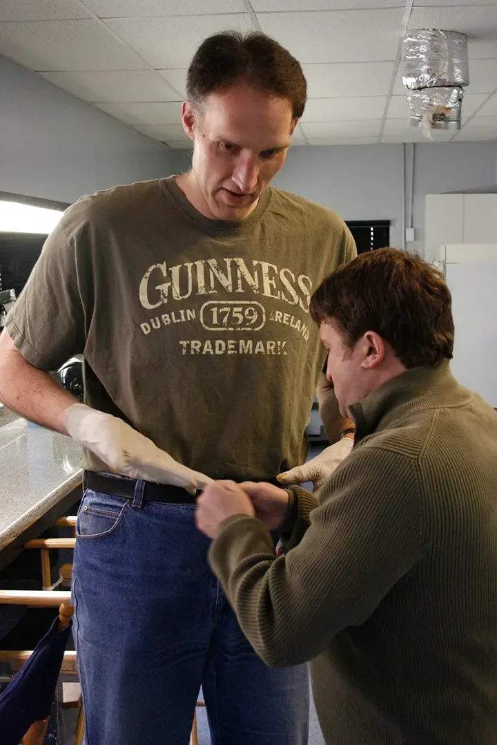 Two men in a workshop measure fabric together, one standing and one kneeling.