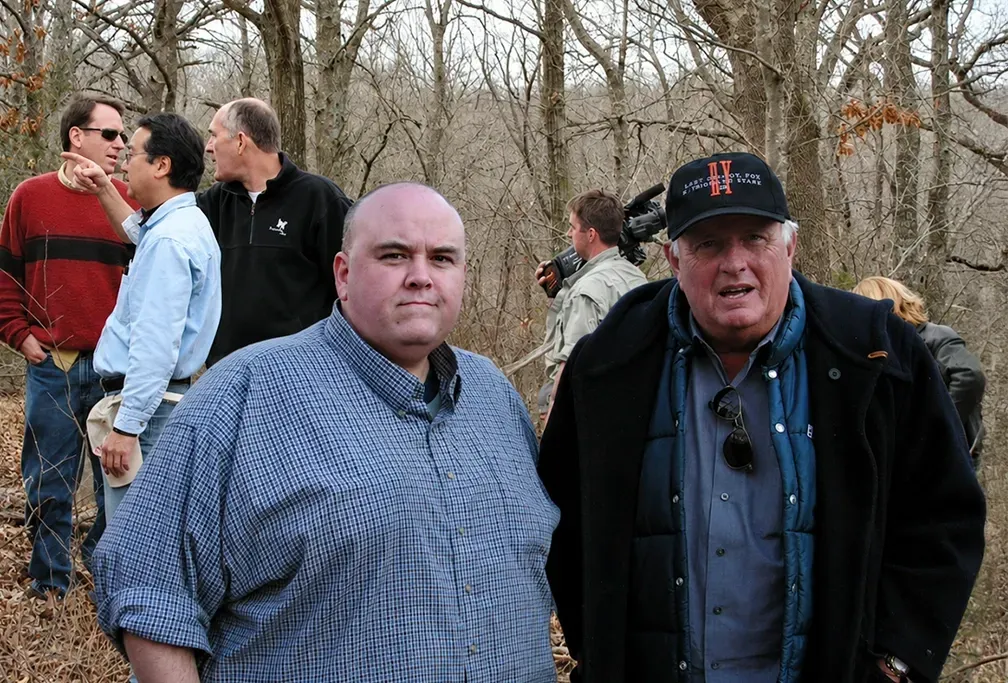 Two men standing outdoors in a wooded area, one in a blue checkered shirt and one in a black jacket and cap.