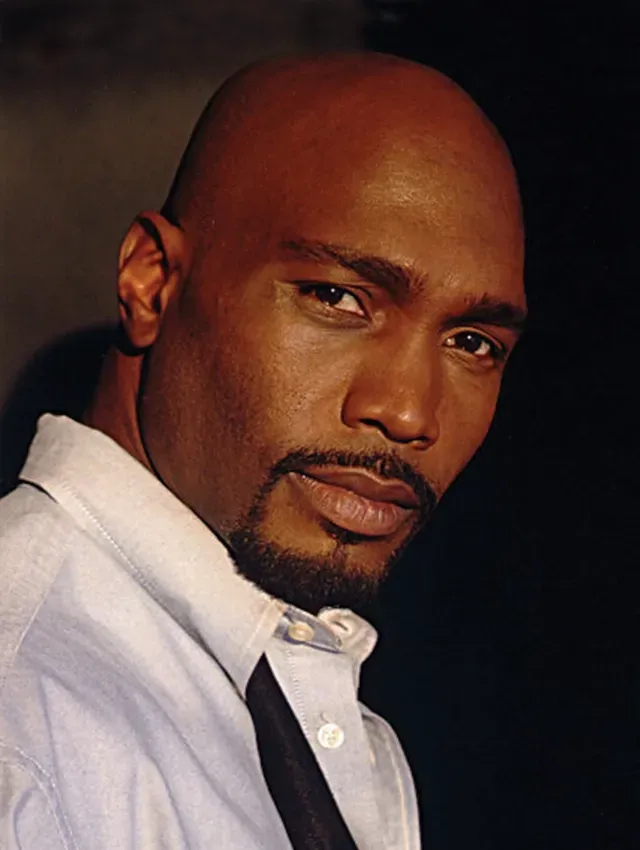 Portrait of a bald man with a goatee in a white shirt and black vest, looking at the camera against a dark background