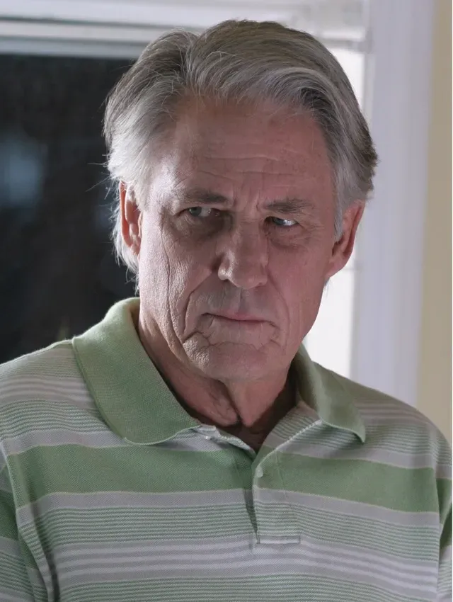 Older man in a green striped polo shirt, looking seriously at the camera indoors.