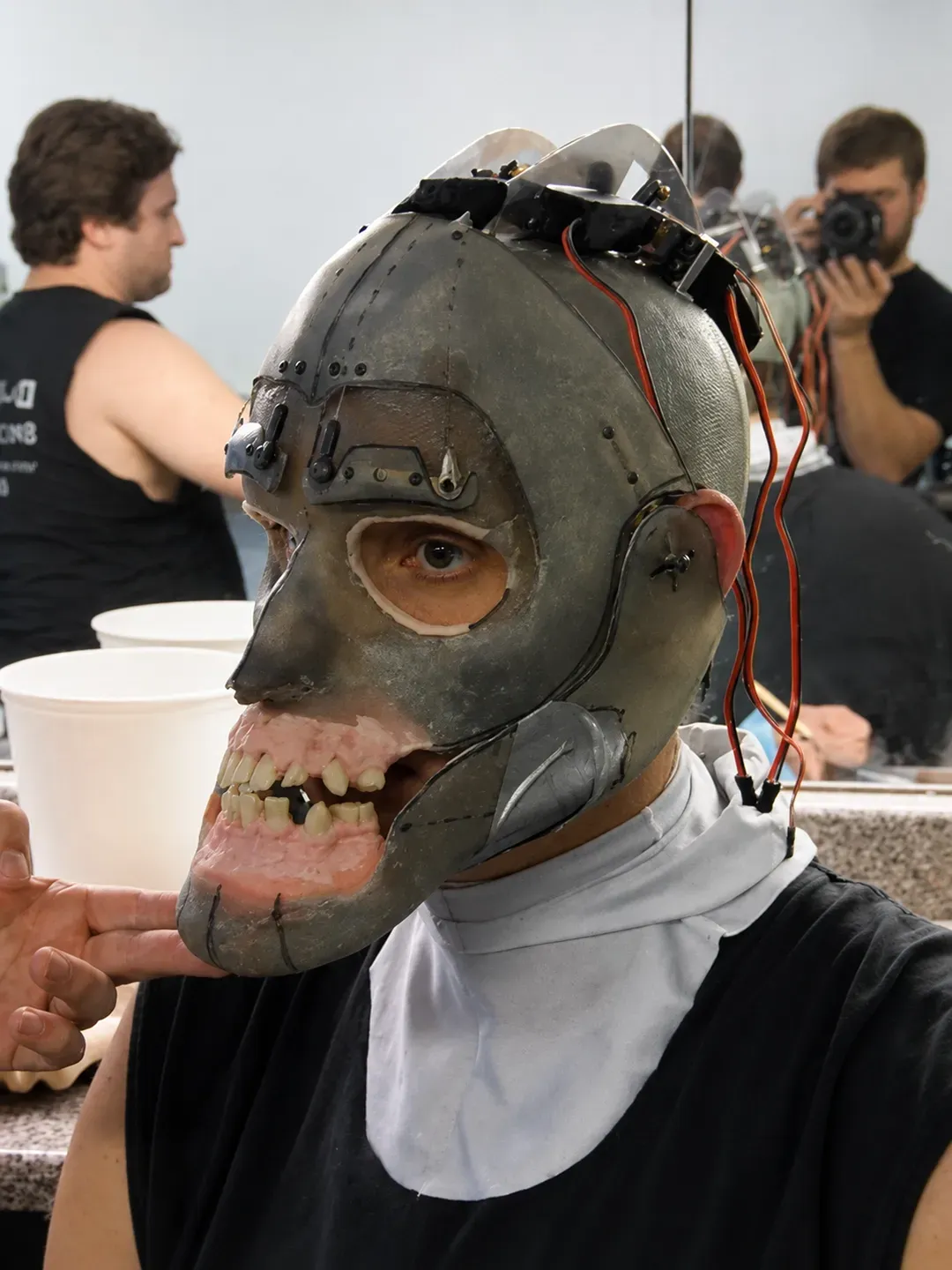 Person wearing a detailed metal face mask at a workshop, with photographers and participants in the background