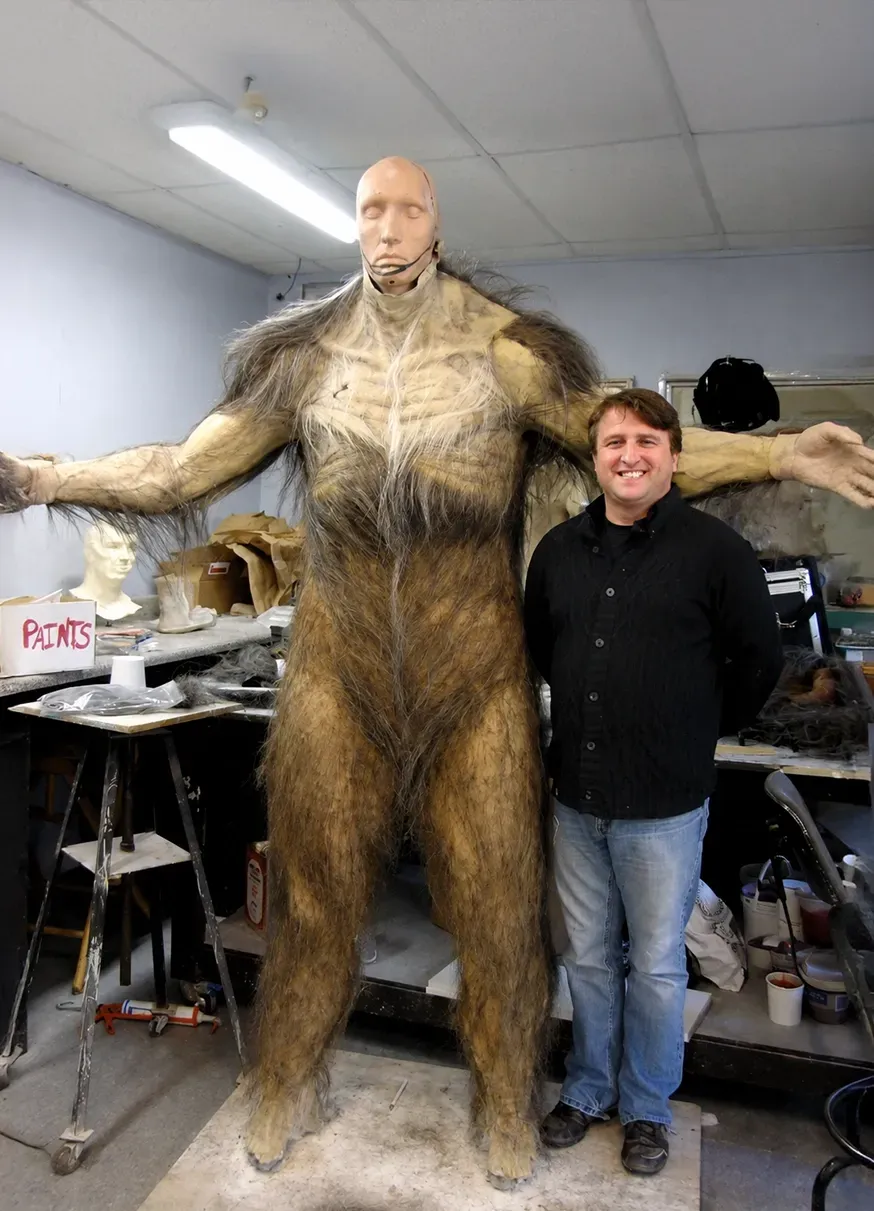 Man standing beside a tall, furry creature costume with arms outstretched in a cluttered workshop