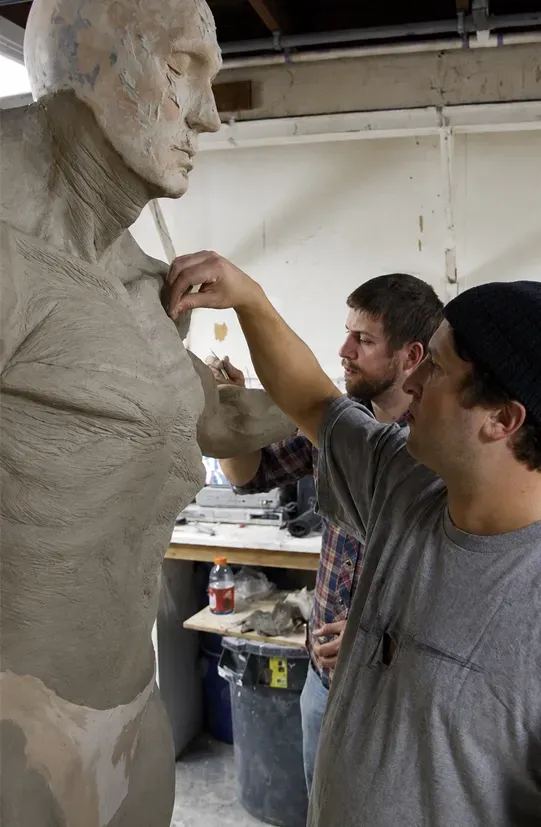 Two people sculpting a clay bust in a studio, one adjusting details on the chest.