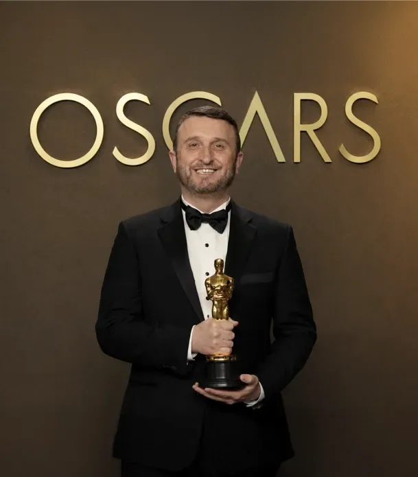 Man in tuxedo holding Oscar statue in front of Oscars backdrop