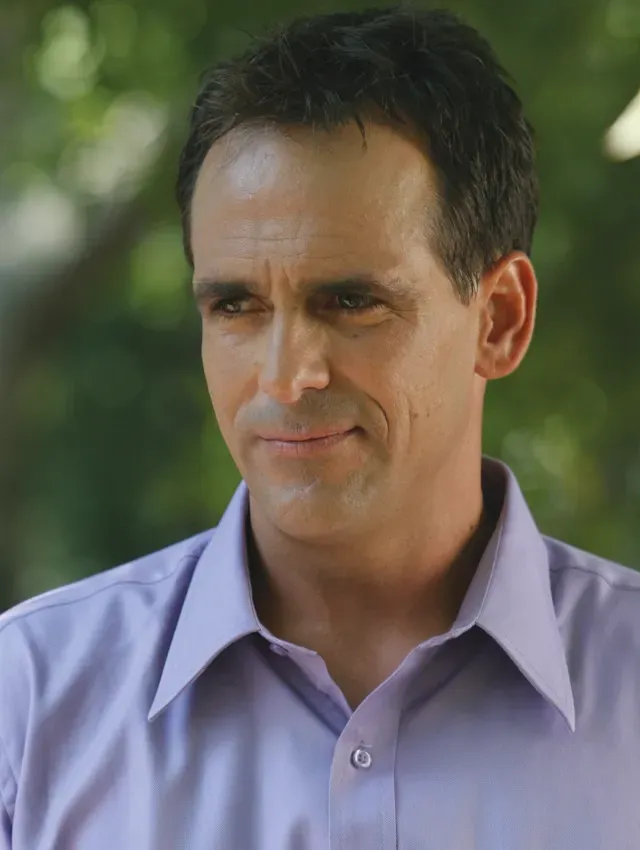 Man in a light purple shirt outdoors, with a neutral expression against a blurred green background