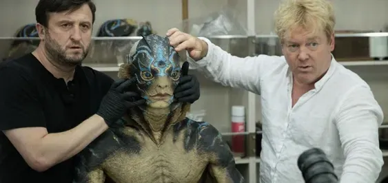 Two men in black and white shirts adjust a blue-costumed creature on a film set