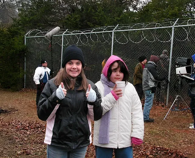Two people in winter jackets and hats give thumbs up outdoors by a fence and group behind them.
