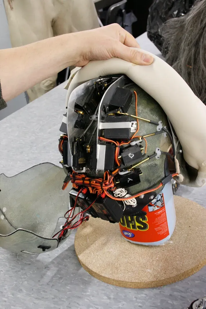 Wired homemade helmet-like device held by a hand, with exposed electronics and red wires on a table.