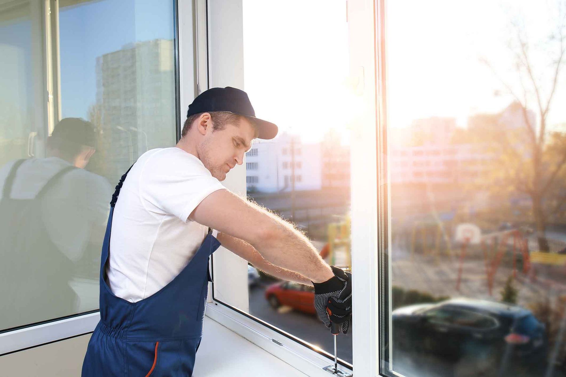 Man in overalls installing a window, using a screwdriver, indoors near a sunny view.