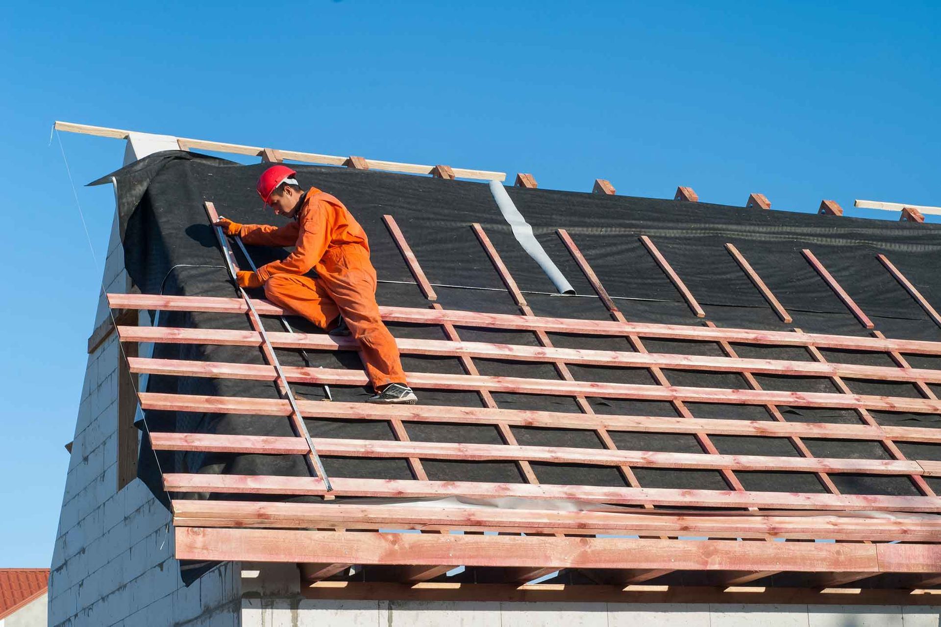 Roofer in orange jumpsuit works on a roof, securing boards with a tool on a sunny day.