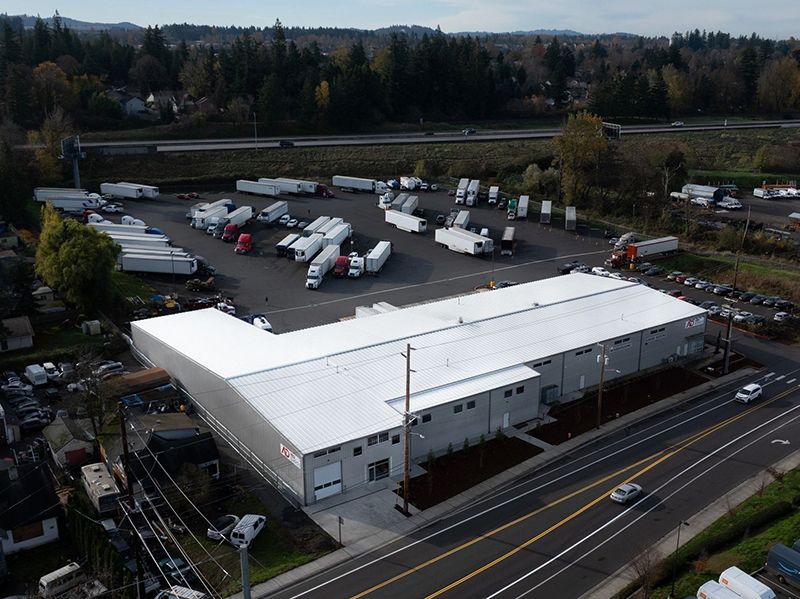 Aerial view of a white warehouse with parked trucks in an industrial yard beside a road.