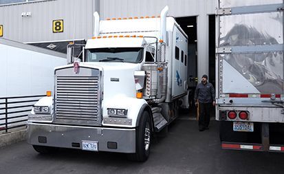 White semi truck parked in a loading dock beside another trailer, with a worker standing nearby