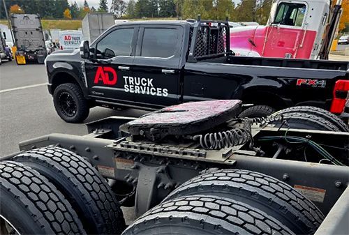 Black AD Truck Services truck with visible fifth-wheel hitch, seen from the rear at a work yard