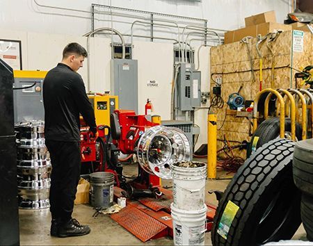 Man inspecting machinery in a workshop with tires, tools, and stacked wheel rims