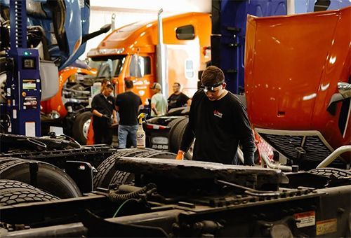 Mechanic inspecting a truck chassis in a busy auto repair shop, with orange semi-trucks in the background