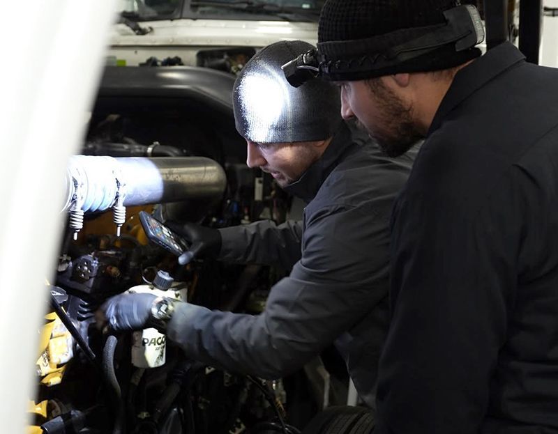 Two technicians in dark clothing inspect engine equipment with a flashlight in a cramped mechanical space.