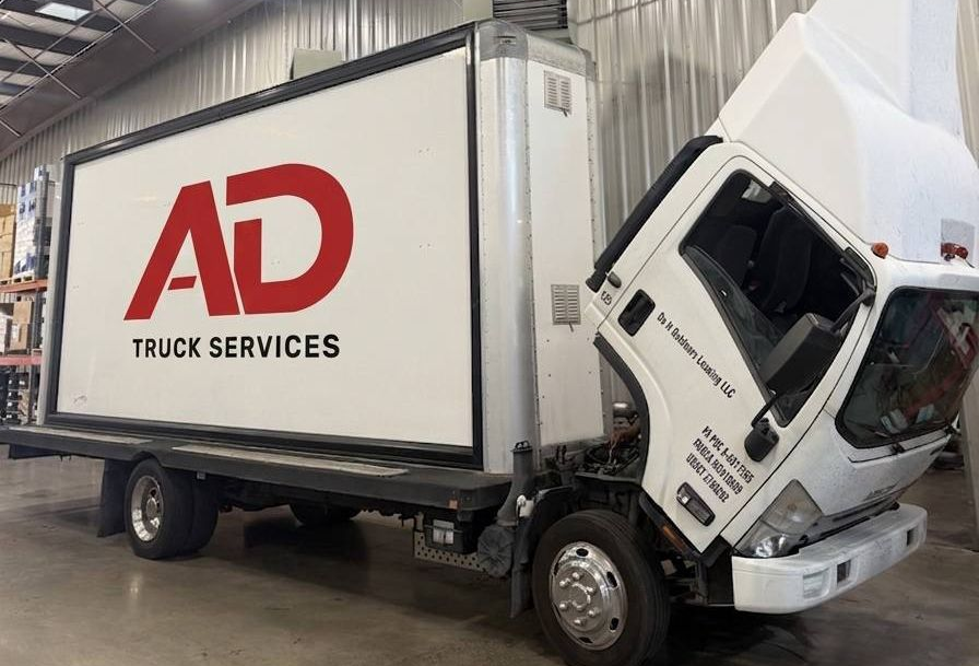 White AD Truck Services box truck tipped forward inside a warehouse, cab crumpled against the floor.