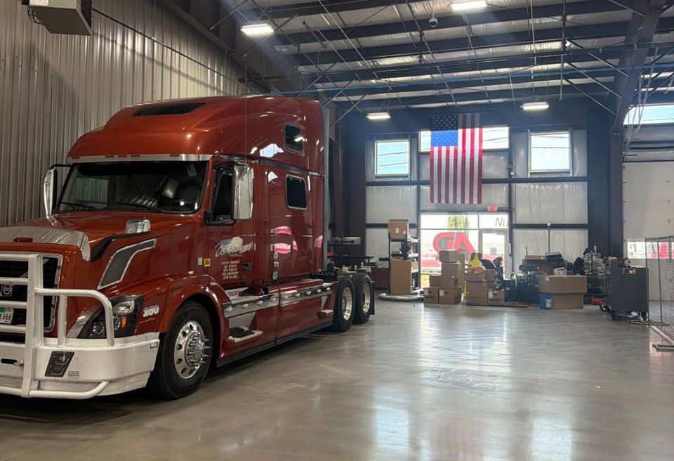 Red semi-truck parked inside a warehouse with an American flag on the back wall.