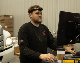 Man at a desk using a desktop computer, wearing a headband and black shirt, with a coffee cup nearby.