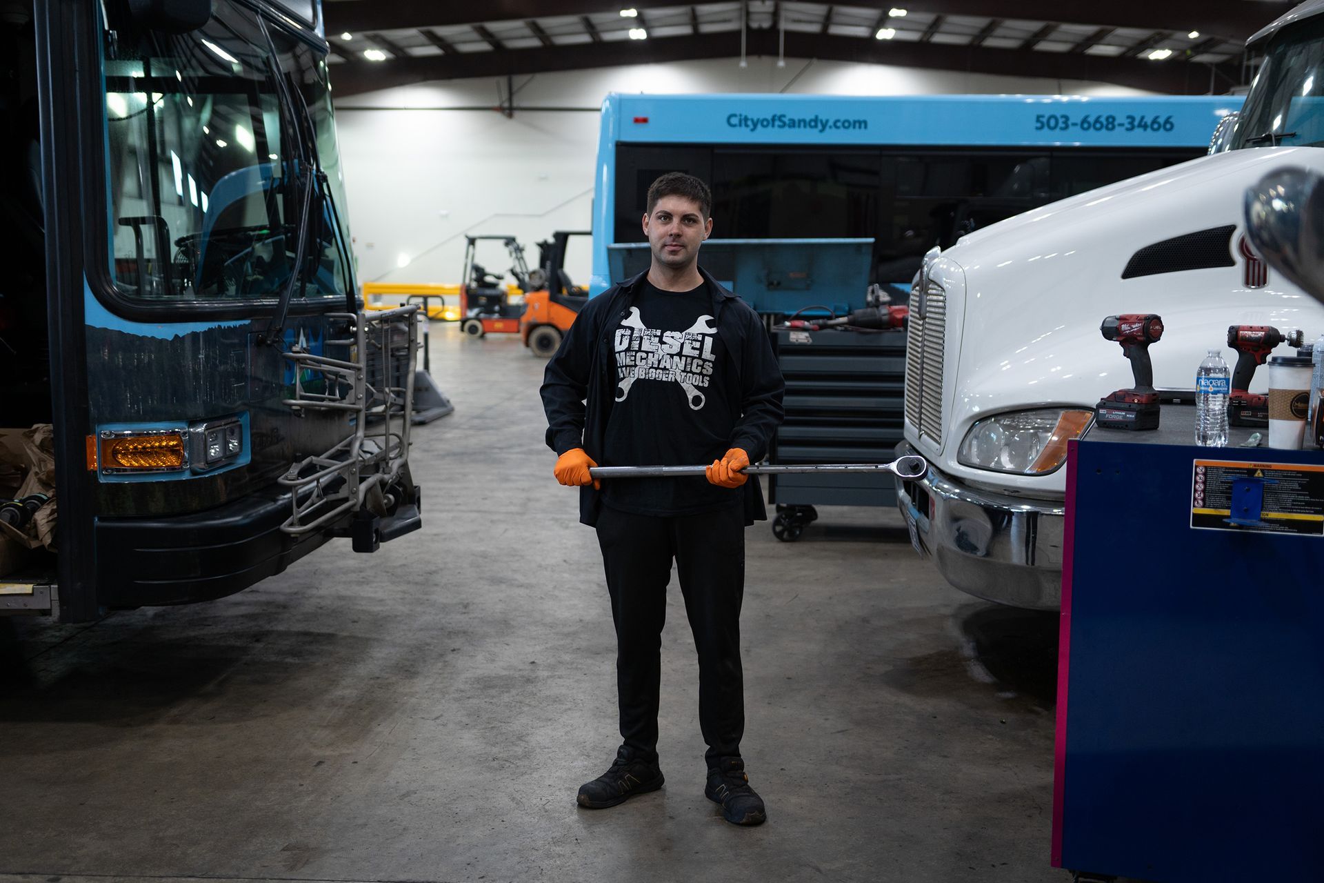Person standing in an auto shop holding a tool between buses and a white truck