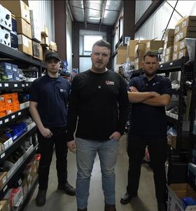 Three men stand in a warehouse aisle surrounded by stacked boxes and shelves of parts.