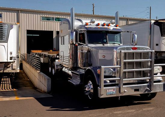 Chrome semi-truck backed to a loading dock at a warehouse, with trailer doors open.